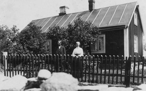 Disgen handledning - Gustav och Berta vid staketet framför sitt hus där stora syrenbuskar skymmer fönstren.
