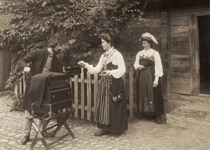 Disgen handlening - En positivhalare tar emot ett mynt av en folkdräktsklädd kvinna. Skansens vårfest 1905. Nordiska Museet.