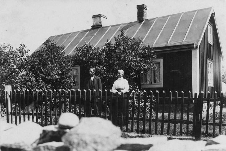 Disgen handledning - Gustav och Berta vid staketet framför sitt hus där stora syrenbuskar skymmer fönstren.